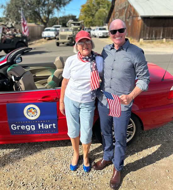 Asm. Hart and Gabi Robbins in front of parade car, with barn and cars in background