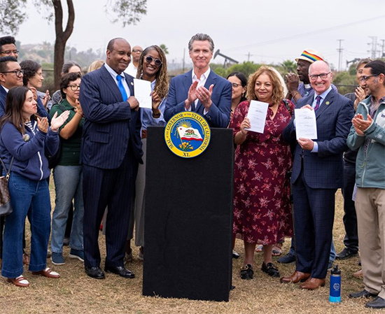 Gov. Newsom at podium, clapping as Asm. Hart and other hold up signed documents