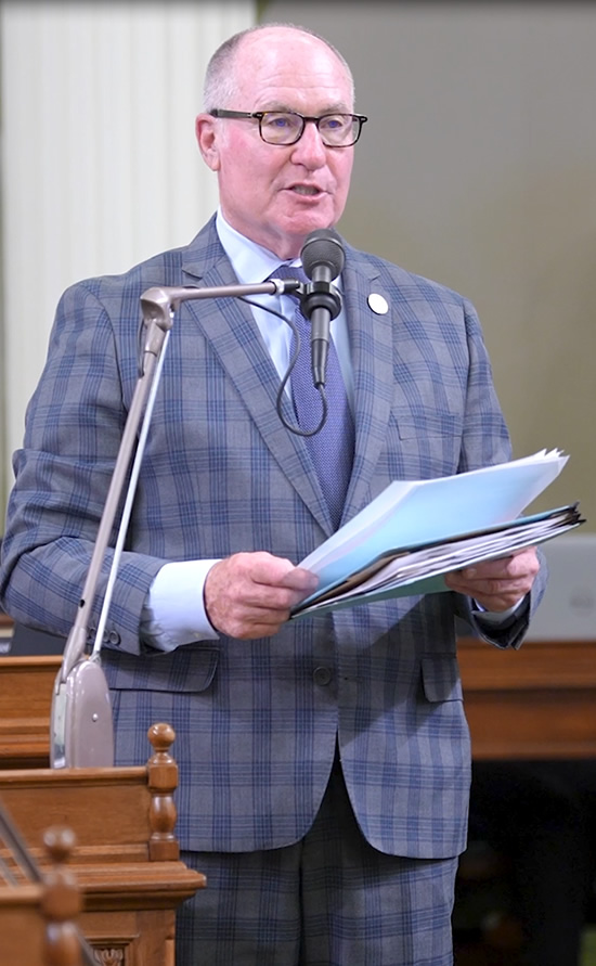Asm. Hart standing at microphone, speaking and holding documents