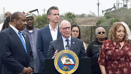 Asm. Hart at podium, speaking, as Gov. Newsom and others listen on behind him