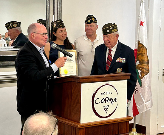 Asm. Hart holding microphone, speaking and standing at podium with three veterans