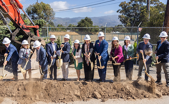 Asm. Hart and others at dirt pile with hard hats on, using shovels to dig