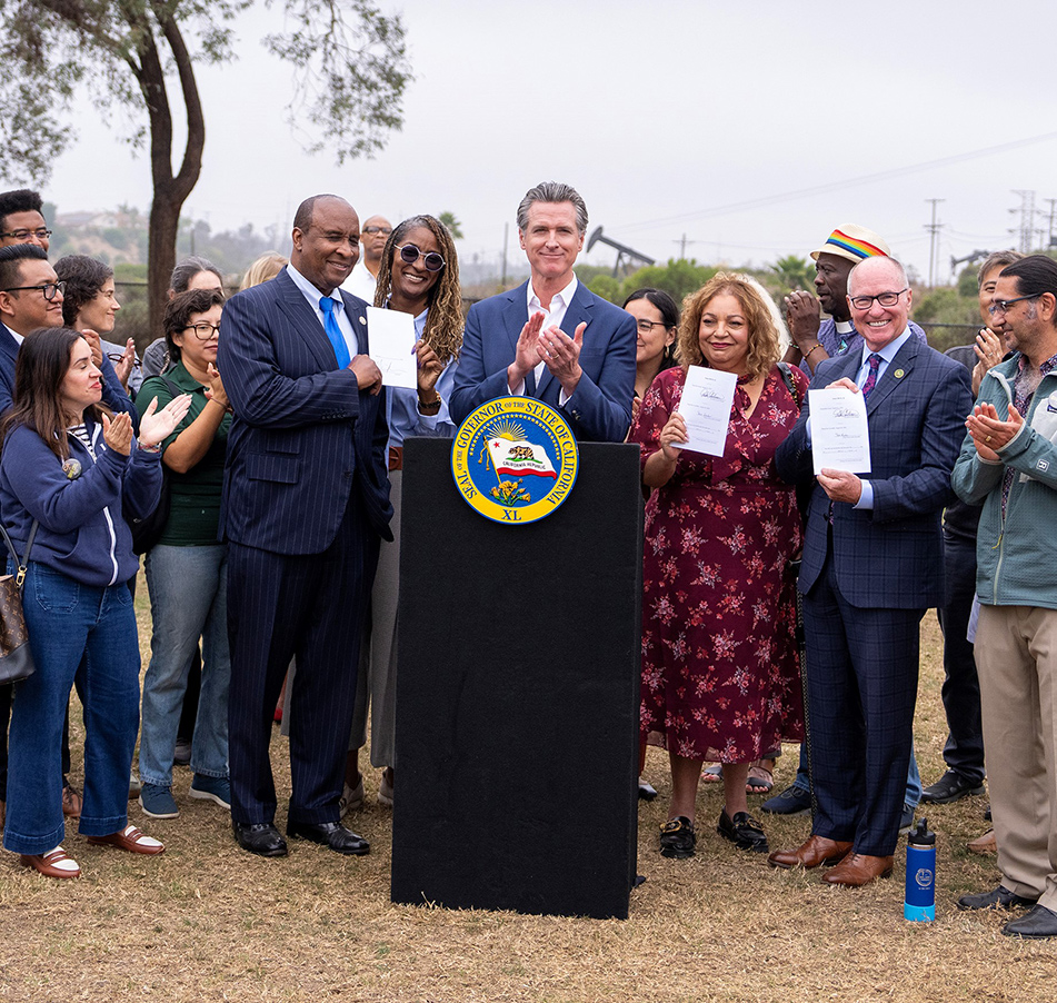 Gov. Newsom at podium, clapping along with other as Asm. Hart and other hold up signed documents 