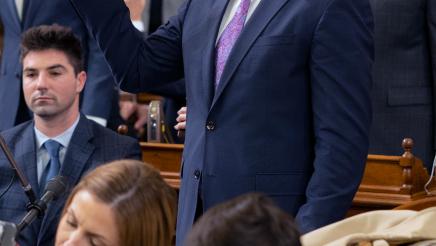 Asm. Hart standing, holding up right hand, with other members in background