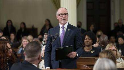 Asm. Hart standing, holding binder and speaking into microphone as other members listen on