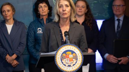 Guest speaker at podium, speaking, while Asm. Hart and other guest speakers listen on
