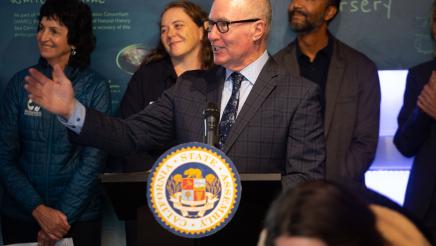 Asm. Hart at podium, speaking and holding up an outstretched arm to one side of the room